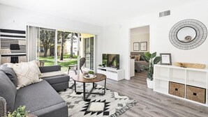 Uma sala de estar moderna com um sofá cinzento, uma mesa de centro e uma TV. Há uma janela ampla com vista para um jardim.