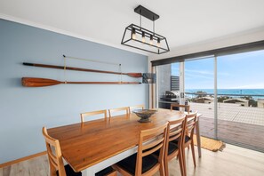 A dining area with a wooden table and chairs, a wall-mounted television, and a view of the ocean through large glass doors. - Pier Vista (Lorne)