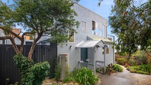 A two-story house with a grey exterior, a front porch with a white railing, and a small garden with various plants and flowers. - Pier Vista (Lorne)