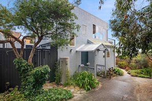 A two-story house with a grey exterior, a front porch with a white railing, and a small garden with various plants and flowers. - Pier Vista (Lorne)