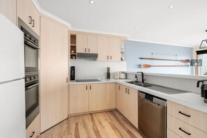 A modern kitchen with wooden cabinets, a built-in oven, a dishwasher, and a countertop with a sink. - Pier Vista (Lorne)