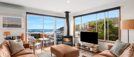 A modern living room with a large TV, leather sofas, a fireplace, and a view of the ocean.