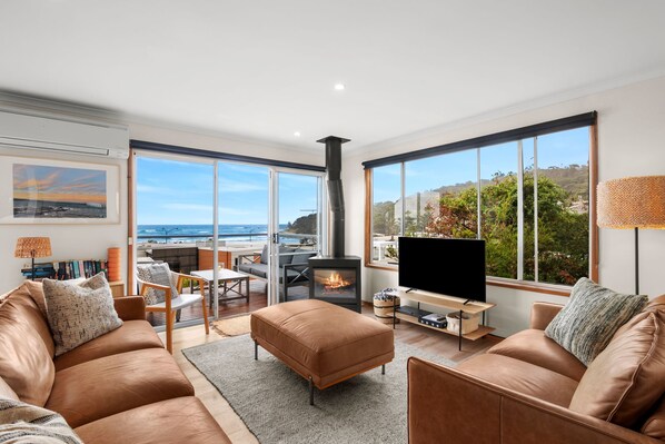 A modern living room with a large TV, leather sofas, a fireplace, and a view of the ocean.