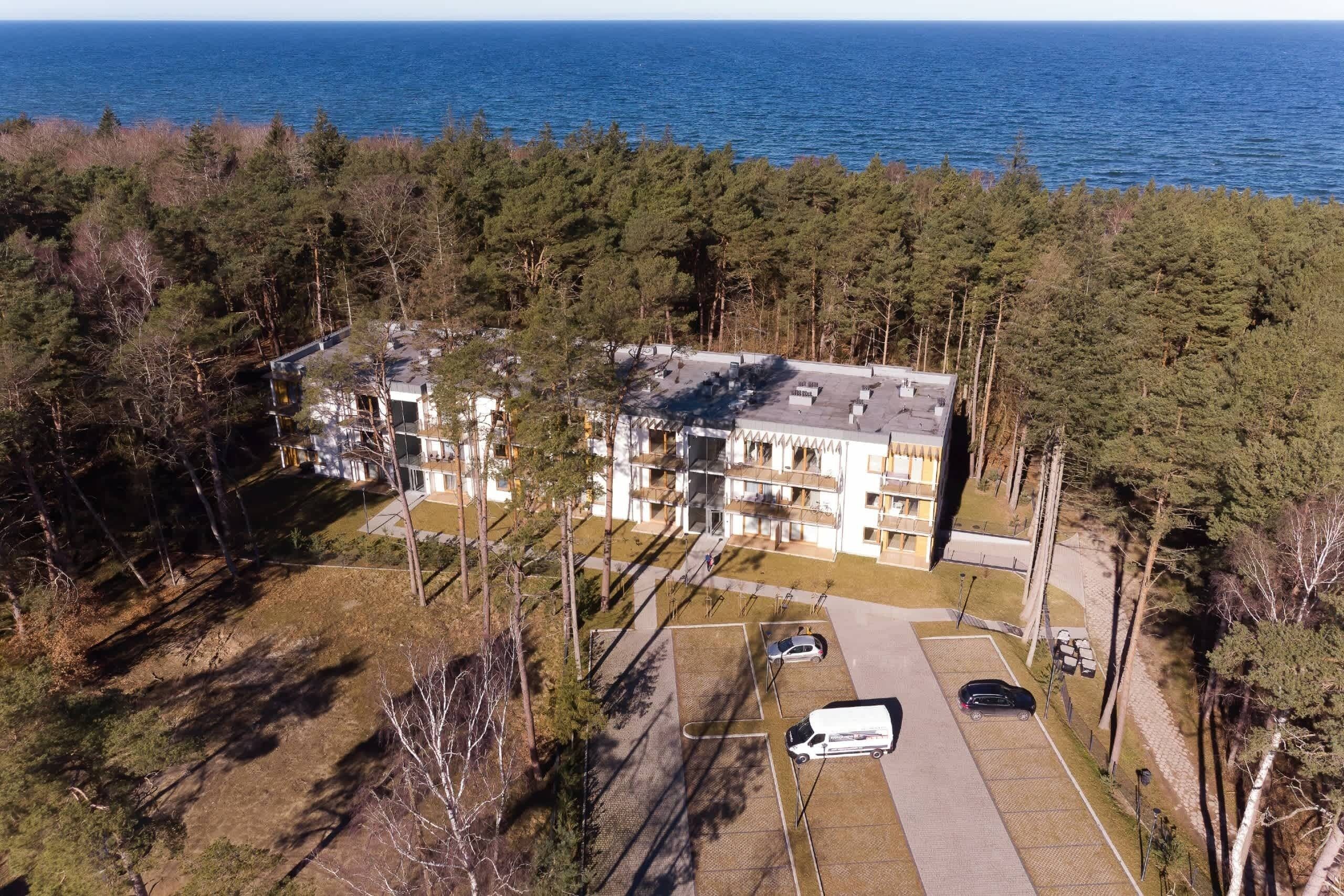 An aerial view of a multi-story building with a parking lot, surrounded by dense forest and overlooking a body of water.