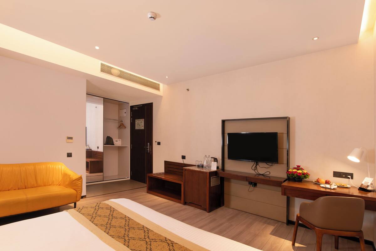 A hotel room with a sofa, a desk with a lamp, a TV, and a small table with flowers and fruit.