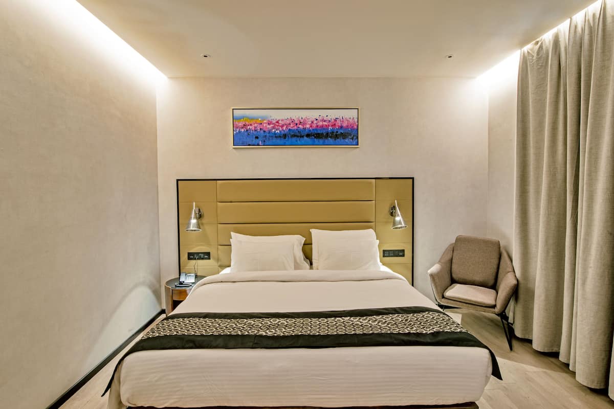 A hotel room with a large bed, a headboard, a chair, a nightstand, a lamp, and a painting on the wall.