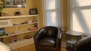 Un salon avec un fauteuil en cuir marron, une étagère blanche contenant des livres et des objets décoratifs, un tableau encadré au mur et une fenêtre avec des stores.