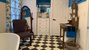 Une salle de bain avec un carrelage à motifs, une chaise en bois, une baignoire blanche, une coiffeuse avec un miroir et une porte donnant sur une autre pièce.