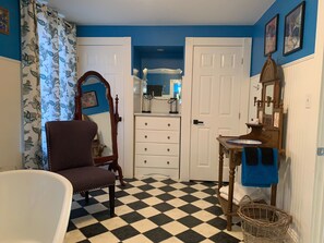 A bathroom with a checkered floor, a wooden chair, a white bathtub, a vanity with a mirror, and a door leading to another room.