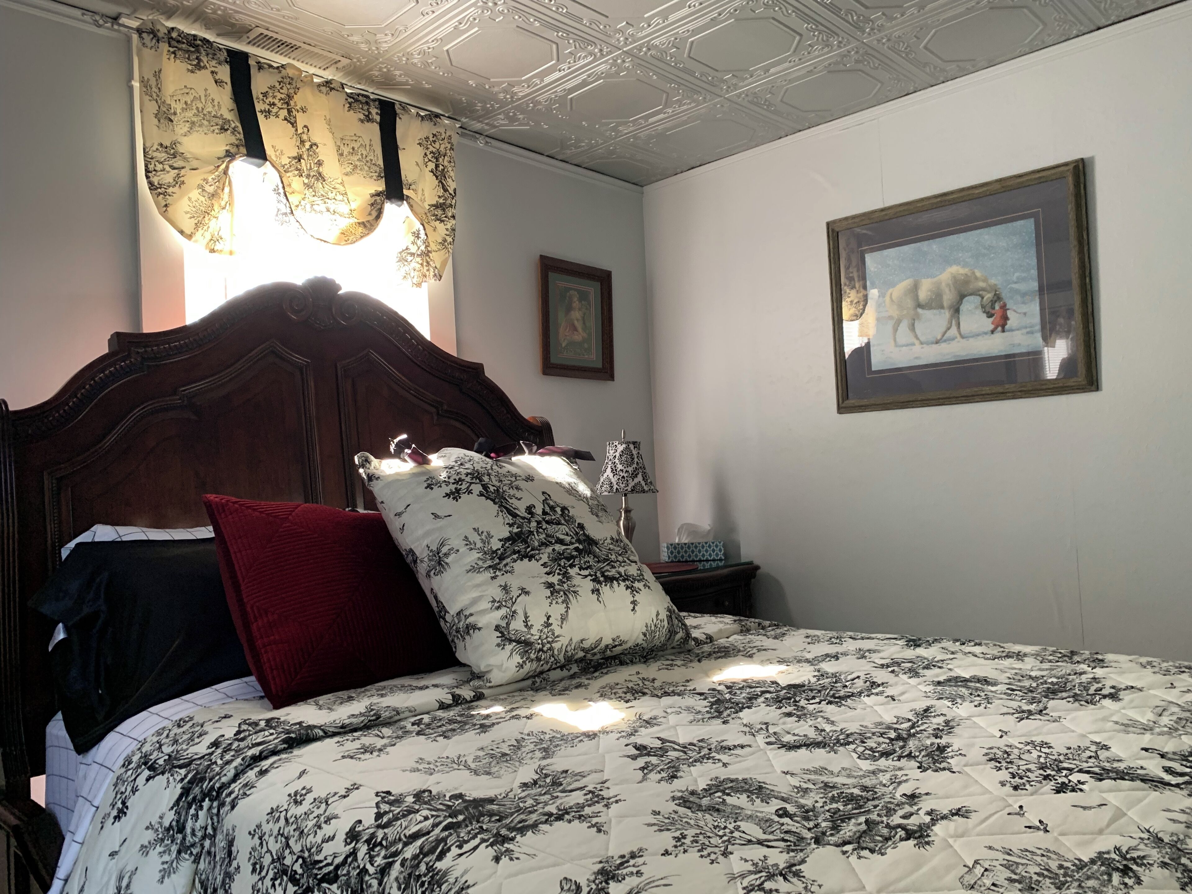 A bedroom with a large bed, floral bedding, a wooden headboard, a nightstand with a lamp, and a framed picture on the wall.