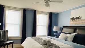 A bedroom with a large bed, a chair, a ceiling fan, and a painting on the wall.