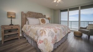 Een slaapkamer met een groot bed, een houten nachtkastje, een plafondventilator en uitzicht op de oceaan vanaf een balkon.