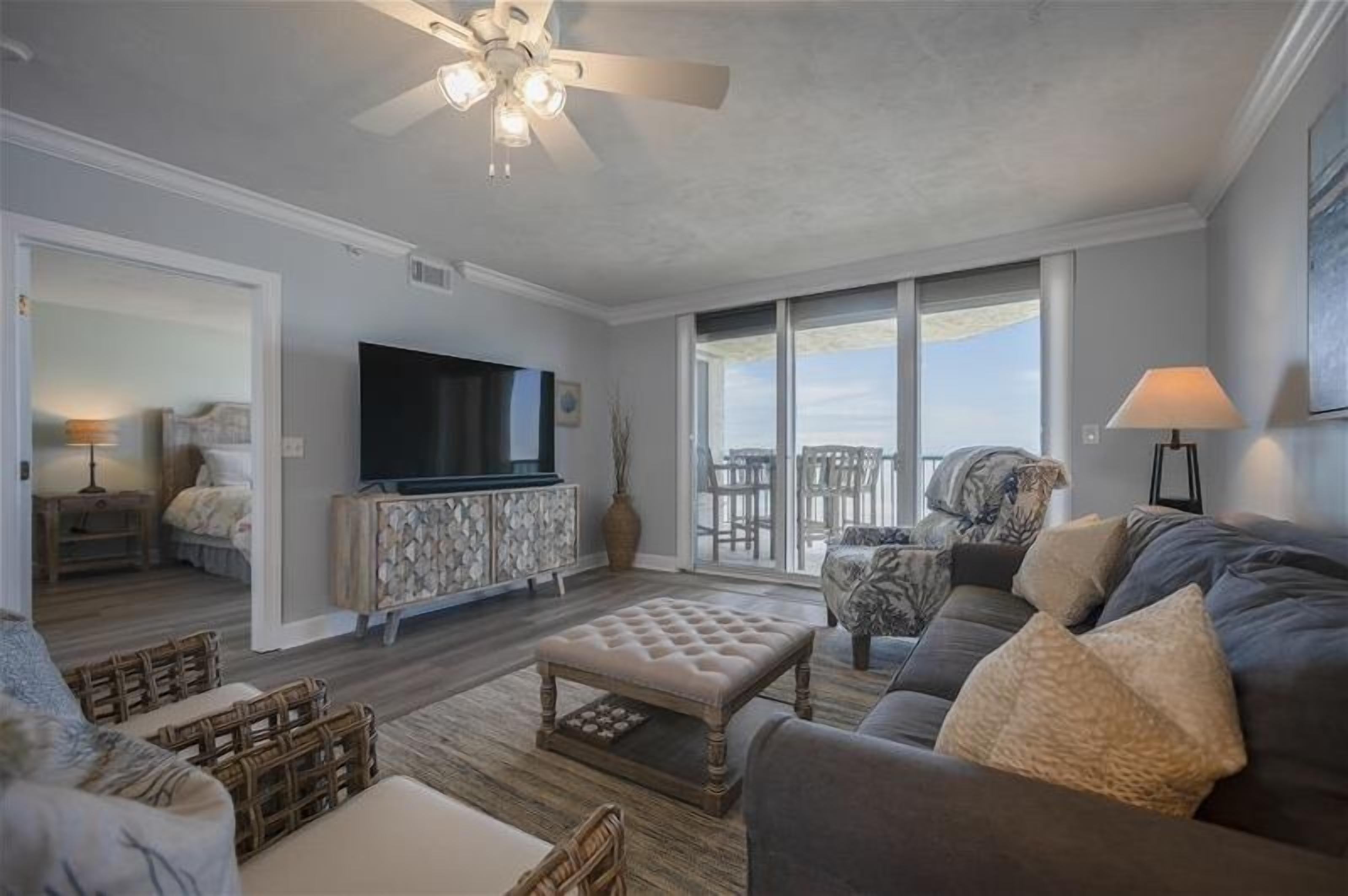 A spacious living room with a large TV, comfortable seating, and a balcony with a view.