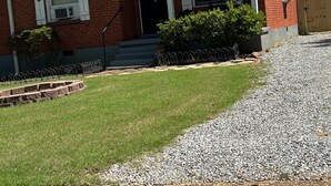 Una casa de ladrillo de un piso con un porche delantero, el número '220' en la puerta, y un pequeño jardín frontal con un sendero de grava y césped.