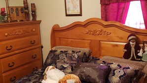 A bedroom with a wooden headboard, patterned bedding, a dresser with drawers, a framed picture on the wall, and a window with curtains. - Just Like Home (Memphis)