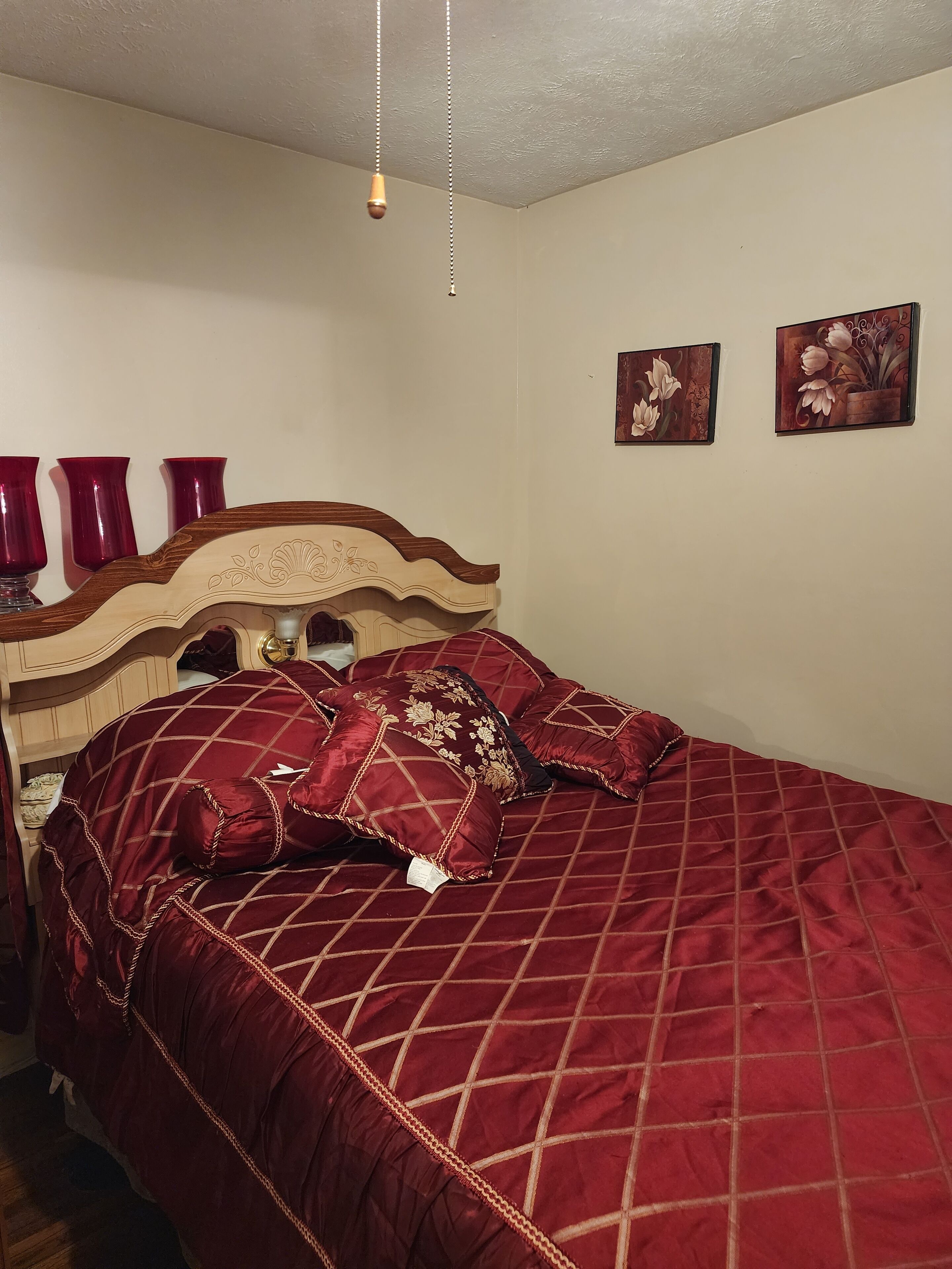 A neatly made bed with a red quilt and matching pillows, a wooden headboard, and two wall-mounted floral paintings.