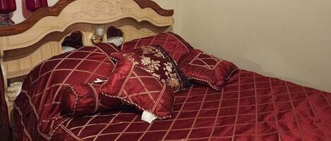 A neatly made bed with a red quilt and matching pillows, a wooden headboard, and two wall-mounted floral paintings.