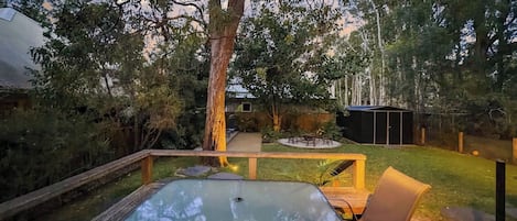An outdoor deck with a glass-top table and chairs, surrounded by a wooden deck and a grassy area with trees.