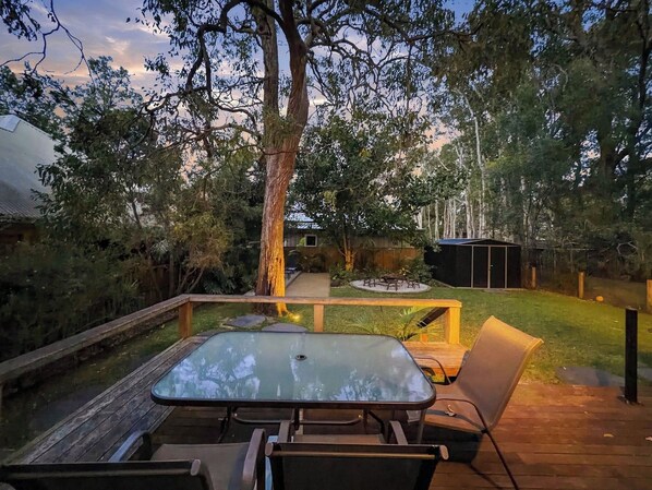 An outdoor deck with a glass-top table and chairs, surrounded by a wooden deck and a grassy area with trees.