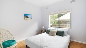 Ein Schlafzimmer mit einem großen Bett, einem Korbsessel, einem Fenster mit Blick auf ein Haus und einem Gemälde an der Wand.