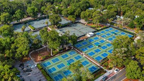Vue aérienne d’un complexe sportif comprenant plusieurs courts de tennis, entourés d’arbres et de zones de stationnement.