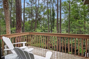 A wooden deck with white Adirondack chairs and a railing overlooking a forested area.
