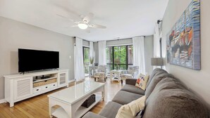 A modern living room with a flat-screen TV, white furniture, a ceiling fan, and large windows offering a view of greenery.