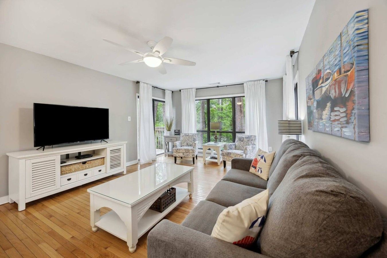 A living room with a flat-screen TV, a white coffee table, a grey sofa, and a ceiling fan.