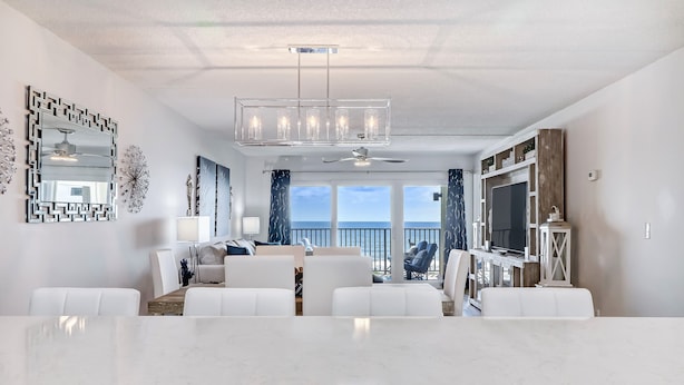 Un salon moderne avec une grande table blanche, des canapés blancs, un ventilateur de plafond et une vue sur l’océan.