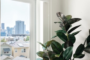 Una habitación con un ventanal grande que da a un paisaje urbano, una planta en maceta y una pared blanca.