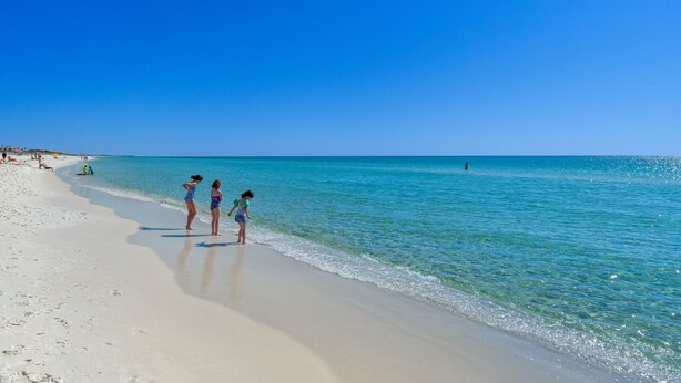 A beach with clear blue water and a few people standing on the sand.