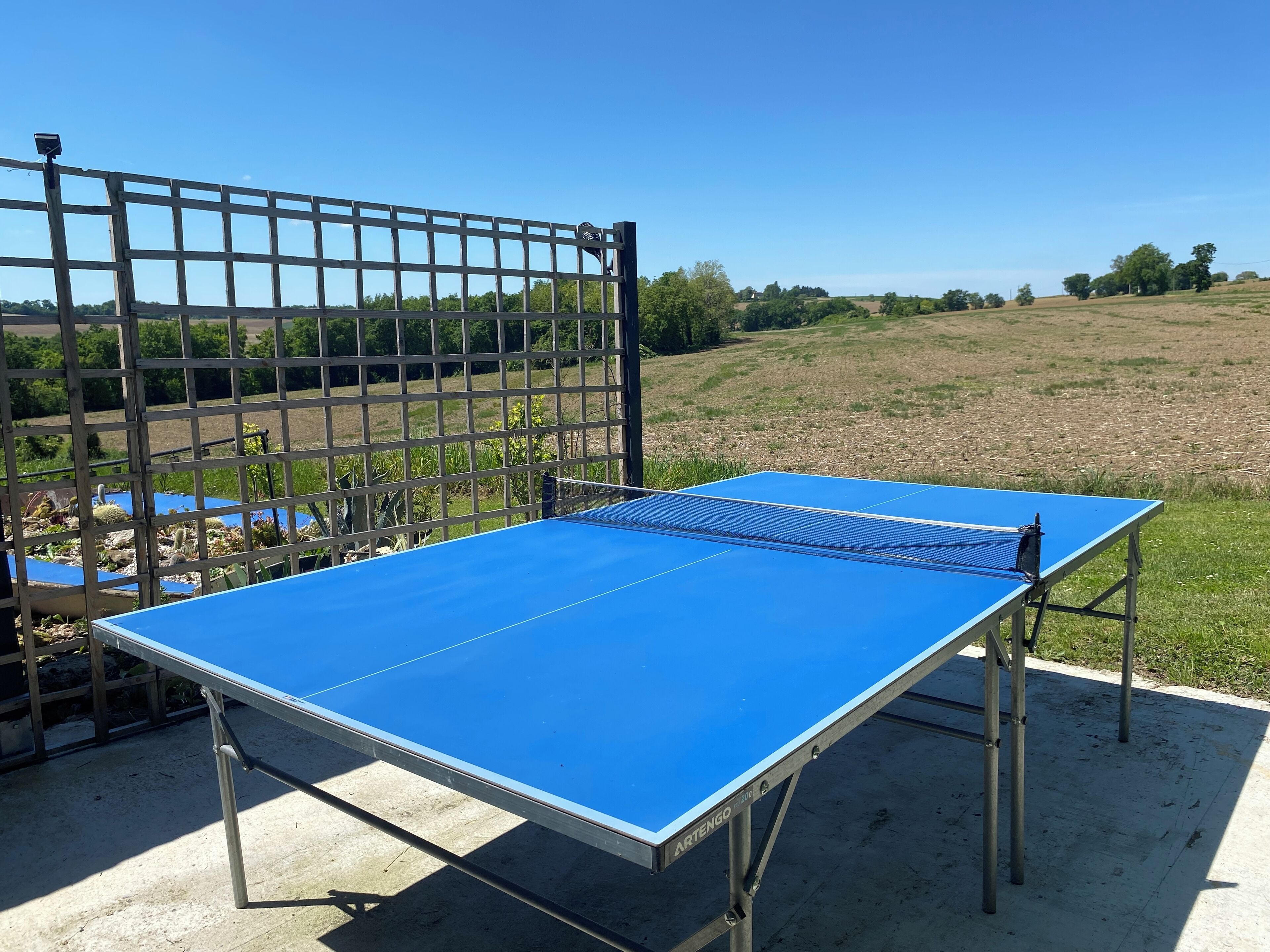 A blue table tennis table with metal legs, set up outdoors with a metal fence in the background.