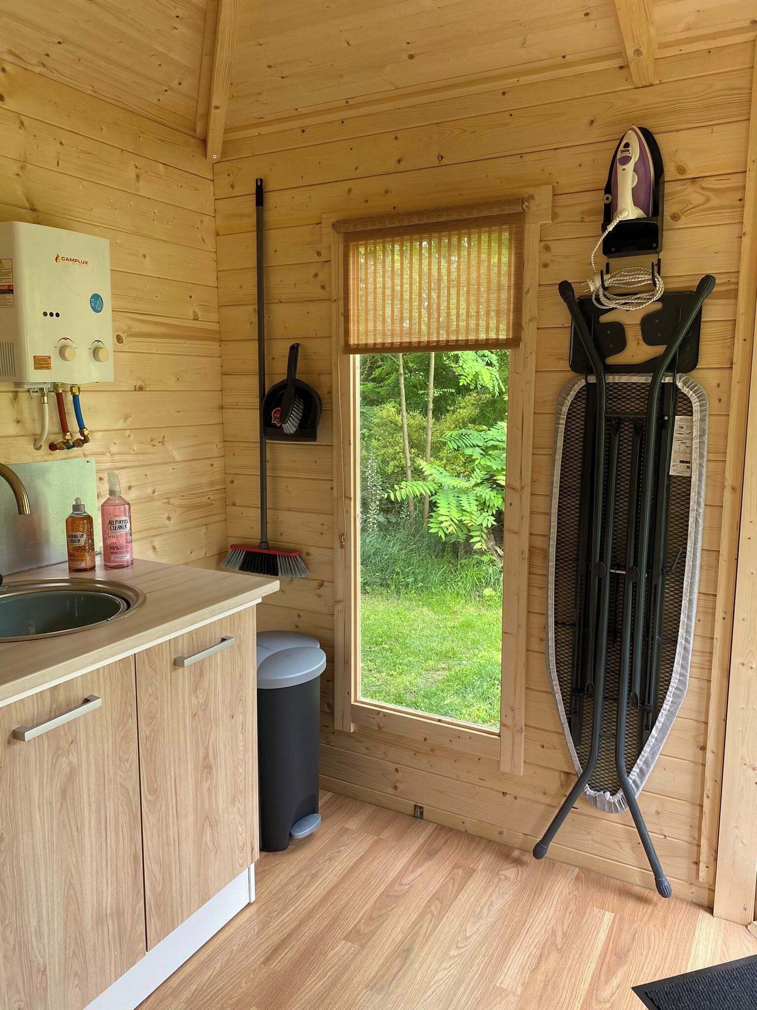 En stuga med träpanel på insidan, med ett köksområde som inkluderar en diskho, en spis och ett fönster med utsikt över grönskan.