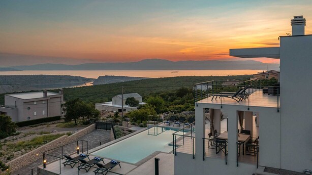 A modern pool area with lounge chairs and a dining set, overlooking a scenic landscape with mountains and a body of water during sunset.