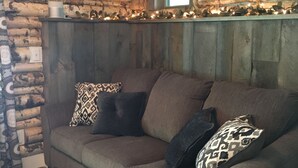 A log cabin interior with a brown sofa, patterned cushions, a clock on the wall, and a window with blinds. - Stonehaven Cottages - "The Birchview Suite Duplex - Lower Level" (Rice Lake)