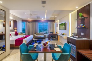 Quarto de hotel com cama, mesa de jantar com cadeiras, televisão e cozinha compacta.