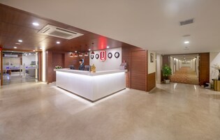 Uma área de recepção de hotel com design moderno, incluindo um balcão de recepção, um relógio fixado na parede e um vaso de planta.