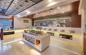 A modern hotel lobby with a central buffet area, a patterned wall, and a view of the cityscape through large windows. - Lords Eco Inn Rajkot Shapar (Kotda Sangani)