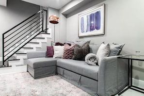 A modern living room with a grey sectional sofa, a black staircase, a floor lamp, and a framed abstract painting on the wall.