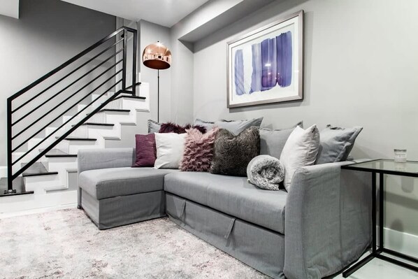 A modern living room with a grey sectional sofa, a black staircase, a floor lamp, and a framed abstract painting on the wall.