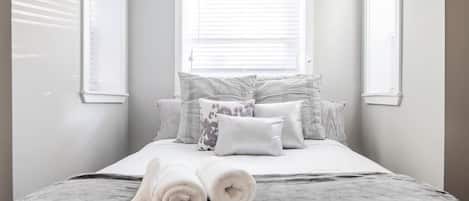 A neatly made bed with rolled towels, a window with blinds, and a plain wall.