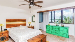 A bedroom with a bed, a green dresser, a ceiling fan, a window with a view, and a wooden bench. - Lagoon Views with Beach Access, Pool and Patio (Cancún Q.R)