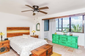 A bedroom with a bed, a green dresser, a ceiling fan, a window with a view, and a wooden bench.