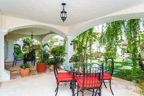 A covered patio with a glass table and red chairs, surrounded by potted plants and columns.