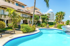 A pool area with lounge chairs, palm trees, and a building with balconies.