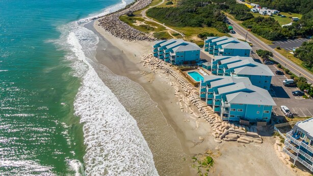 Vista aérea de un hotel frente al mar con varias edificaciones azules, una playa de arena y una vista despejada del océano.