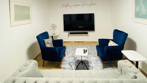 Una sala de estar con un televisor de pantalla plana grande, dos sillones azules, un sofá gris con capitoné y una mesa de centro.