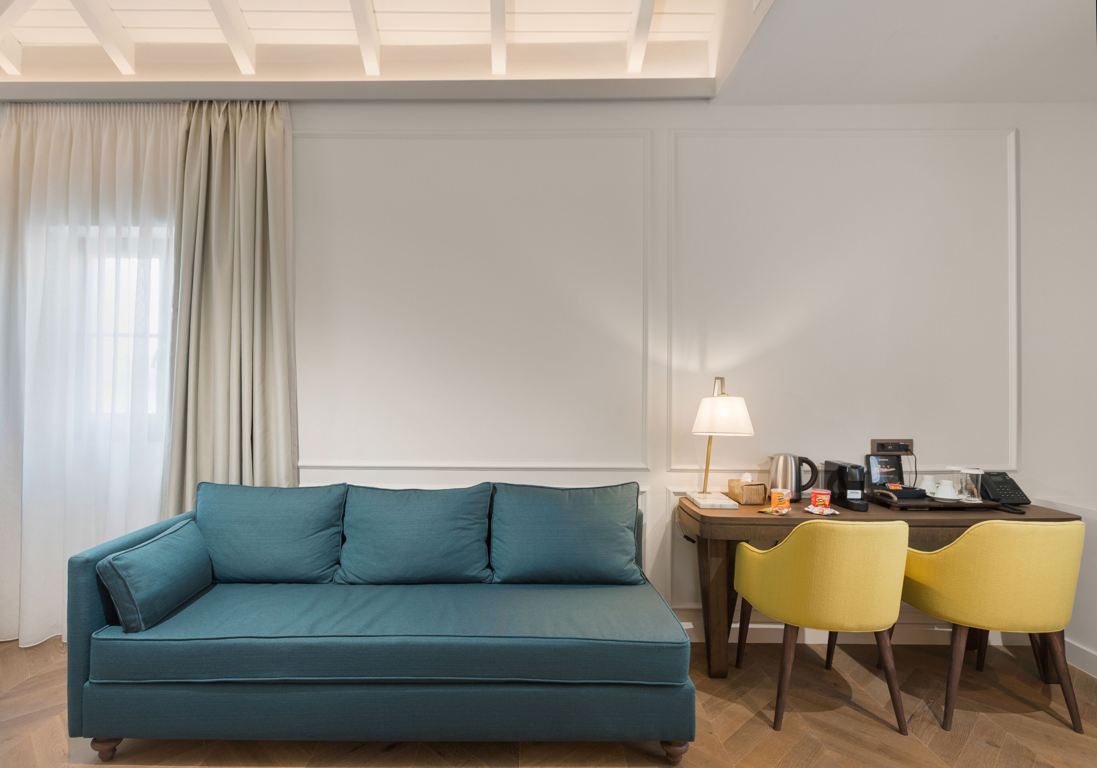 Un canapé de couleur bleu-vert avec des coussins assortis, une table basse en bois, deux chaises jaunes, une lampe et un bureau avec un téléphone et une machine à café.