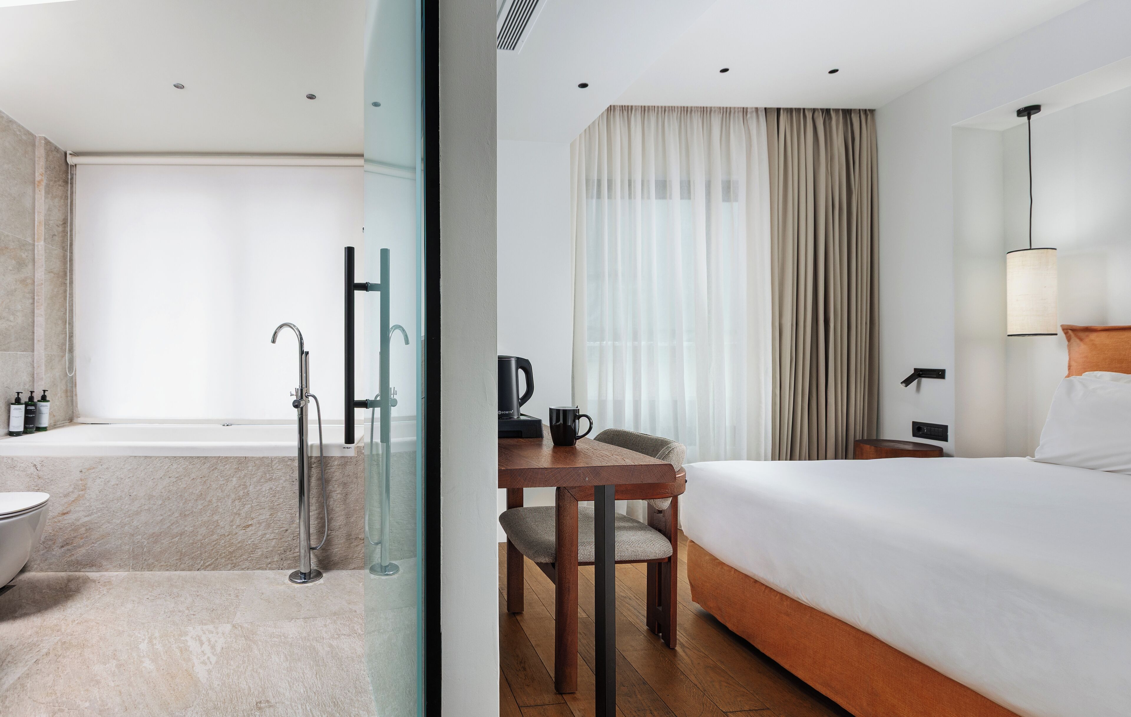 A modern hotel room with a bathtub, a wooden desk with a chair, a bed with white linens, and a large window with sheer curtains.