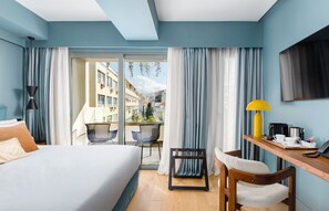 A hotel room with a bed, a desk with a lamp, a chair, and a view of the city through large windows.